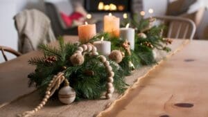 A festive table centerpiece with lit candles, pine branches, wooden beads, ornaments, and dried oranges brings Boho Christmas Decor charm to a burlap runner. A blurred background shows a cozy living room with a fireplace.