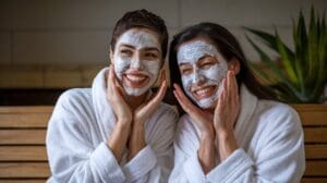 Two women wearing white bathrobes and DIY face masks smile and touch their faces while sitting on a wooden bench. A green plant is visible in the background.