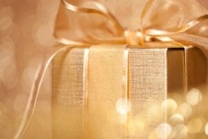 A close-up of a gold gift box with a shiny ribbon and bow, set against a softly blurred background with warm bokeh—perfect inspiration for gifts for the woman who has everything.