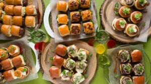 An overhead view of assorted sliders and mini sandwiches on wooden platters, surrounded by various colorful drinks on a green table. The display showcases creative sliders recipes with different toppings, sauces, and vibrant garnishes.