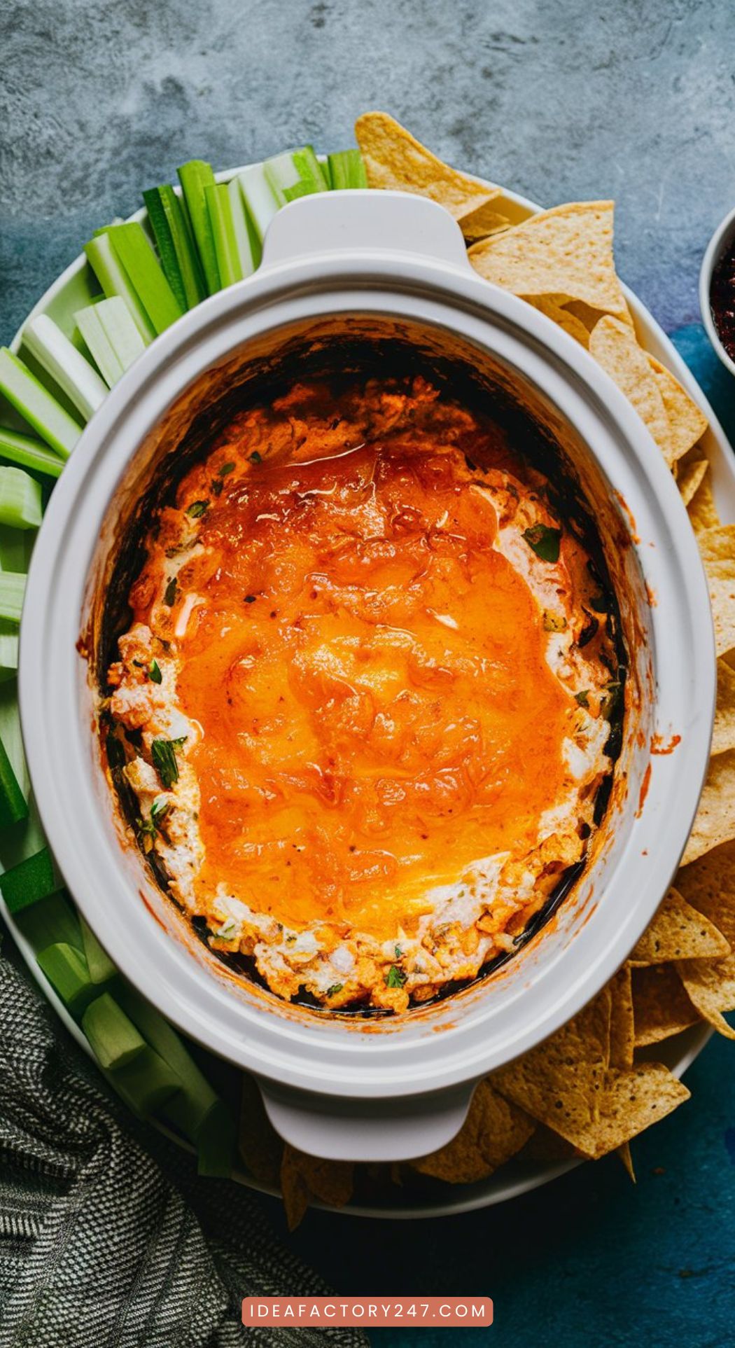 A crockpot filled with creamy buffalo chicken dip, topped with a sprinkle of cheddar cheese, served alongside a platter of celery sticks and tortilla chips.