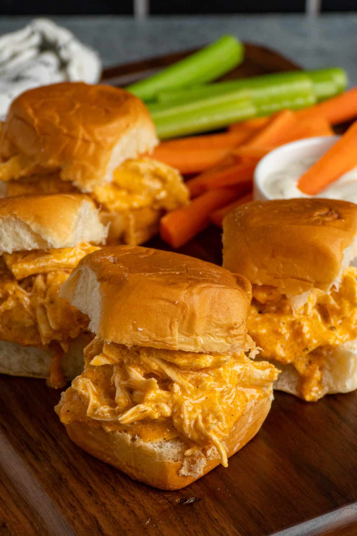 Three buffalo chicken sliders with shredded chicken in orange sauce on soft buns are on a wooden board, served with celery, carrot sticks, and a cup of white dipping sauce—perfect for fans of crockpot appetizer recipes.