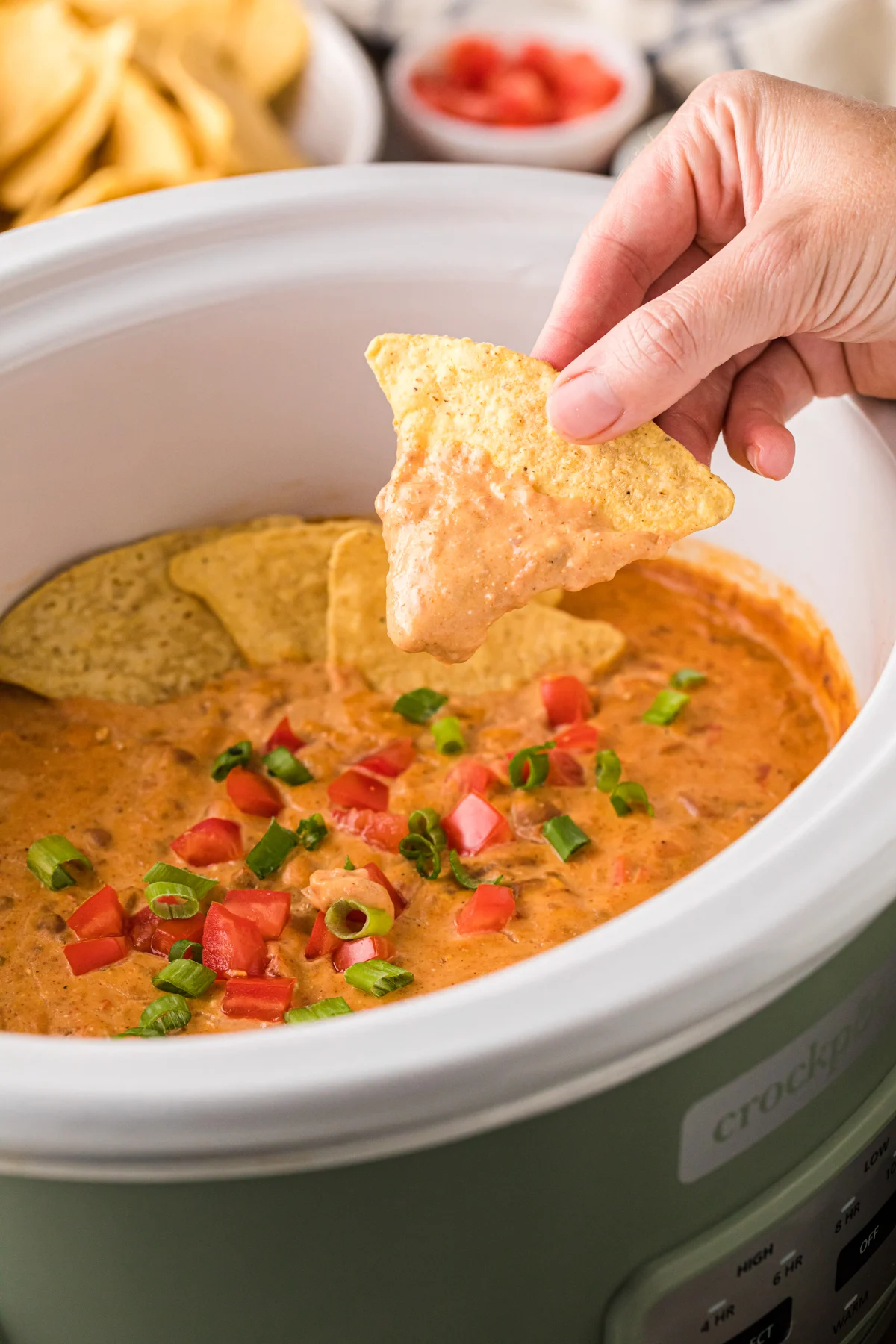 A hand dips a tortilla chip into a crockpot cheese dip topped with chopped tomatoes and green onions—a savory addition to your favorite crockpot appetizer recipes. Other tortilla chips surround the creamy dip.