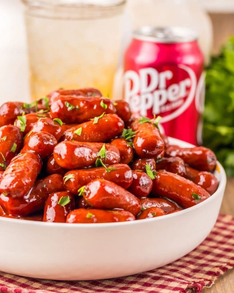 A white bowl filled with glazed cocktail sausages—one of the best crockpot appetizer recipes—garnished with herbs sits on a red and white checkered cloth. In the background, there’s a can of Dr. Pepper and a glass of ice.