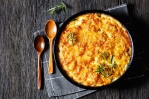 A round baking dish filled with golden, cheesy potato gratin—a classic from easy casserole recipes—garnished with fresh rosemary, sits on a gray checkered cloth next to two wooden spoons on a dark wooden table.