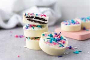 A stack of round Chocolate Dipped Oreos covered in white chocolate and colorful sprinkles, with one cut in half to reveal the classic sandwich cookie inside. Other decorated treats are blurred in the background.