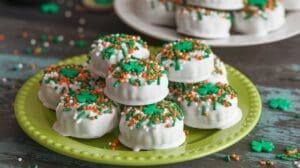 A green plate holds stacked dipped Oreo cookies coated in white icing, topped with green and orange sprinkles and decorated with green shamrock shapes. More cookies are visible on a white plate in the background.