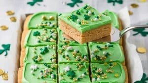 A tray of St. Patrick’s Day Sugar Cookie Bars topped with green icing, festive shamrock and gold bead sprinkles; one bar is lifted with a spatula. Shamrock confetti and gold coins decorate the background.