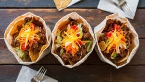 Three paper bowls filled with walking tacos—ground beef, shredded cheese, lettuce, tomatoes, and tortilla chips—sit on a rustic wooden table with napkins and forks nearby.