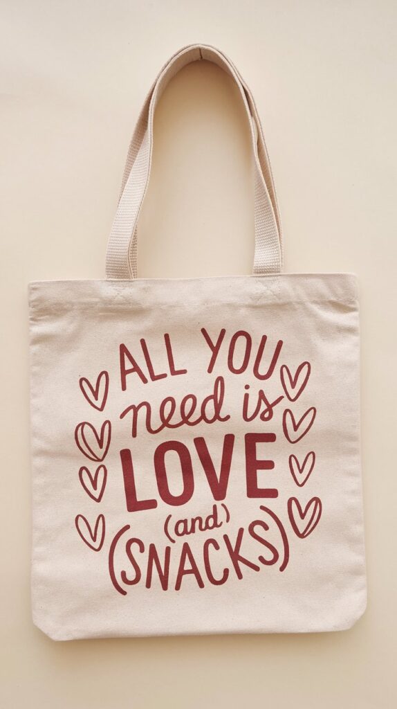 A canvas tote bag with the phrase “All You Need Is Love (and Snacks)” in playful red lettering, surrounded by hand-drawn hearts.

Why They Sell:
Tote bags are practical and reusable, making them a popular eco-friendly gift. Adding Valentine’s-themed designs makes them festive and fun.