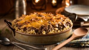 A baked casserole dish filled with noodles, beef, mushrooms, and topped with melted cheddar cheese sits on a rustic wooden table, surrounded by cutlery, plates, and warm ambient lighting.