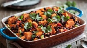 A sweet potato and black bean casserole filled with baked sweet potatoes, black beans, red bell peppers, and celery topped with fresh cilantro sits on a wooden table.