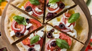 A Whole Wheat Mediterranean Flatbread Pizza topped with tomato, onion, black olives, feta cheese, and fresh basil leaves, served sliced on a wooden board.