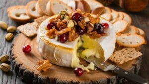 A wheel of baked brie topped with nuts and cranberries sits on a wooden board, surrounded by sliced baguette and crackers. A knife is inserted into the creamy baked brie, ready to be served.