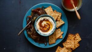A blue plate with crackers, caramelized onions, and a bowl of Caramelized Onion and Goat Cheese Dip topped with honey, next to a spoon and a bowl of honey with a honey dipper on a dark surface.