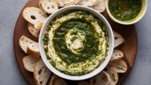 A white bowl of Chimichurri White Bean Dip topped with green herb sauce sits on a round wooden board, surrounded by slices of crusty bread and a small bowl of extra sauce nearby.