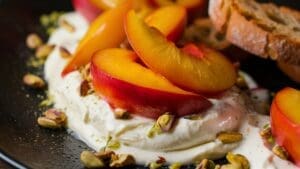 Sliced fresh plums arranged on creamy whipped cheese, inspired by Peach and Burrata Bruschetta Dip, garnished with chopped pistachios, served on a black plate with toasted bread slices in the background.