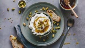 A bowl of creamy yogurt topped with honey and chopped pistachios sits on a plate with slices of bread, creating a delightful spread reminiscent of Peach and Burrata Bruschetta Dip. Nearby are a honey jar, pistachios, and a spoon.