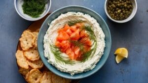 A bowl of creamy dip topped with smoked salmon and fresh dill, surrounded by crispy bread chips, makes this Creamy Smoked Salmon and Dill Spread Recipe perfect. Nearby are bowls of fresh dill, capers, and lemon wedges on a blue surface.