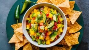 A white bowl filled with vibrant Mango and Avocado Salsa—diced mango, bell pepper, avocado, and herbs—surrounded by tortilla chips and two green chili peppers on a dark green plate.