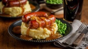 A classic Bangers and Mash plate featuring mashed potatoes topped with sausages, caramelized onions, and rich gravy, served with green peas. A glass of dark beer and utensils rest on a rustic wooden table.