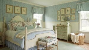 A cozy blue cottagecore bedroom with gingham wallpaper, a cream bed with floral bedding, vintage-style furniture, framed botanical prints on the walls, and sunlight streaming through two windows with matching curtains.