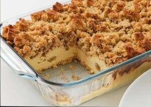 A glass baking dish containing a crumb-topped Cinnamon Roll French Toast Bake with a slice missing, revealing a moist, custard-like interior with apple chunks. Crumbs are scattered on the bottom of the dish.
