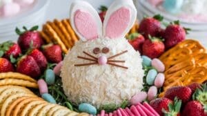 A round easter bunny cheeseball with marshmallow ears, candy whiskers, and a pink nose, surrounded by strawberries, crackers, pretzels, and pastel candy eggs on a platter.
