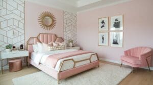 Chic pink bedroom with gold decor, geometric wallpaper, a stylish pink bed, white bedding with pink accents, sunburst mirror, framed art, a pink chair, white rug, and modern nightstands topped with plants.