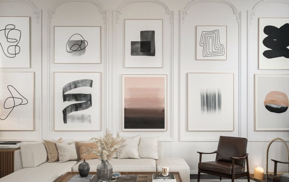 A stylish living room features a white sectional sofa, a brown leather chair, and a wooden coffee table adorned with dried flowers. The wall showcases ten minimalist abstract paintings in black, gray, and soft pink tones, each framed in crisp white.