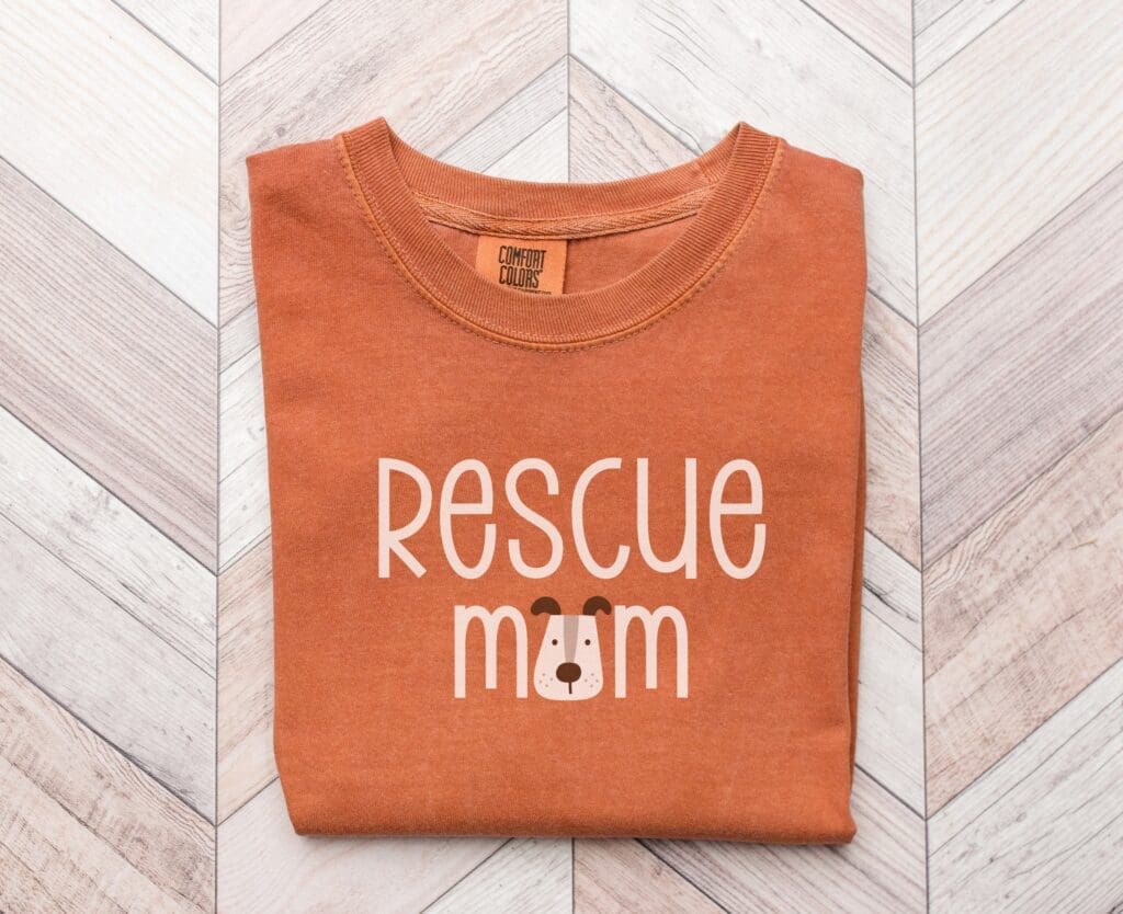 Orange t-shirt featuring the words Rescue Mom with a cute illustration of a dogs face integrated into the design, perfect for gifts. The shirt is neatly folded on a wooden chevron-patterned surface.