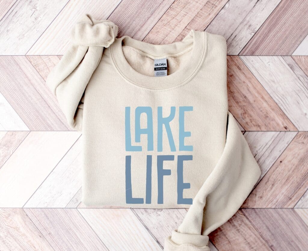 A cozy beige sweatshirt with LAKE LIFE in bold blue letters is neatly folded, making it a perfect gift idea, resting on a light wood herringbone-patterned surface.