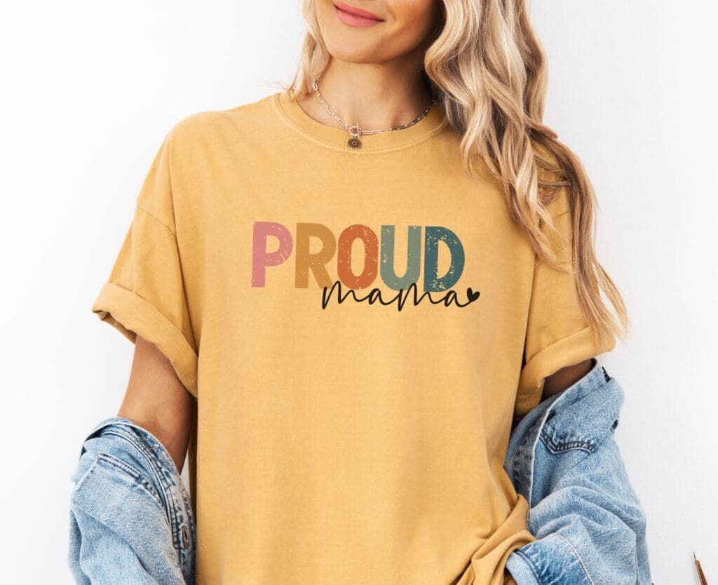 A person with long blonde hair wears a yellow T-shirt with the words Proud Mama in colorful, bold letters, making it an ideal gift for any mom. A denim jacket is draped over their arms against a plain white backdrop.