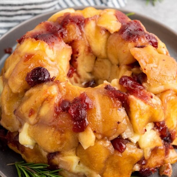 A Brie Pull-Apart Bread ring with golden-brown dough, melted cheese, and chunks of cranberry sauce sits on a gray plate, garnished with fresh rosemary sprigs beside a striped cloth napkin.