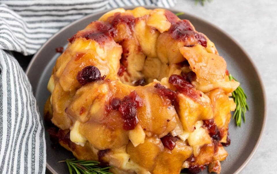 A Brie Pull-Apart Bread ring with golden-brown dough, melted cheese, and chunks of cranberry sauce sits on a gray plate, garnished with fresh rosemary sprigs beside a striped cloth napkin.