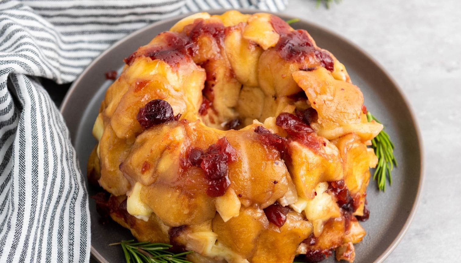 A Brie Pull-Apart Bread ring with golden-brown dough, melted cheese, and chunks of cranberry sauce sits on a gray plate, garnished with fresh rosemary sprigs beside a striped cloth napkin.