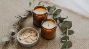 Two brown glass candles and a small bowl of white wax flakes are arranged on a burlap surface, surrounded by eucalyptus leaves—perfect inspiration for your next eucalyptus DIY candle project.