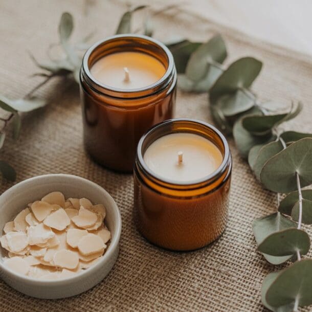 Two brown glass candles and a small bowl of white wax flakes are arranged on a burlap surface, surrounded by eucalyptus leaves—perfect inspiration for your next eucalyptus DIY candle project.