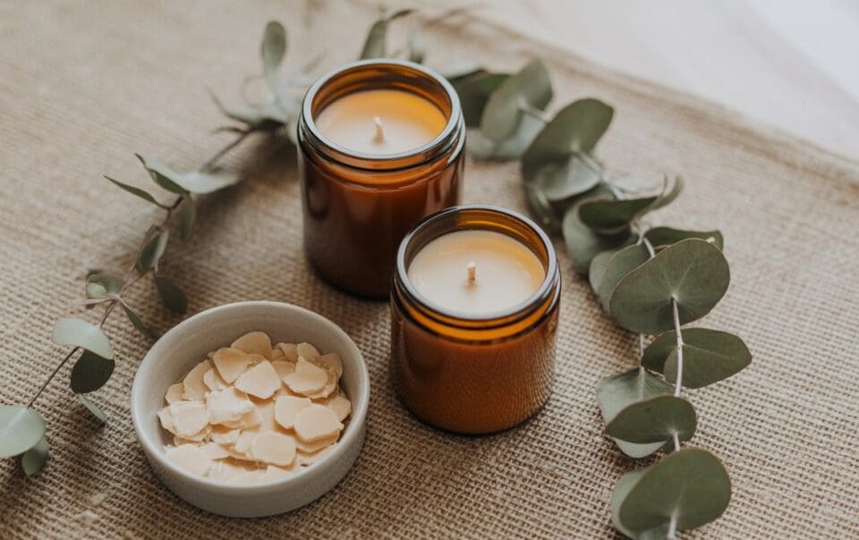 Two brown glass candles and a small bowl of white wax flakes are arranged on a burlap surface, surrounded by eucalyptus leaves—perfect inspiration for your next eucalyptus DIY candle project.