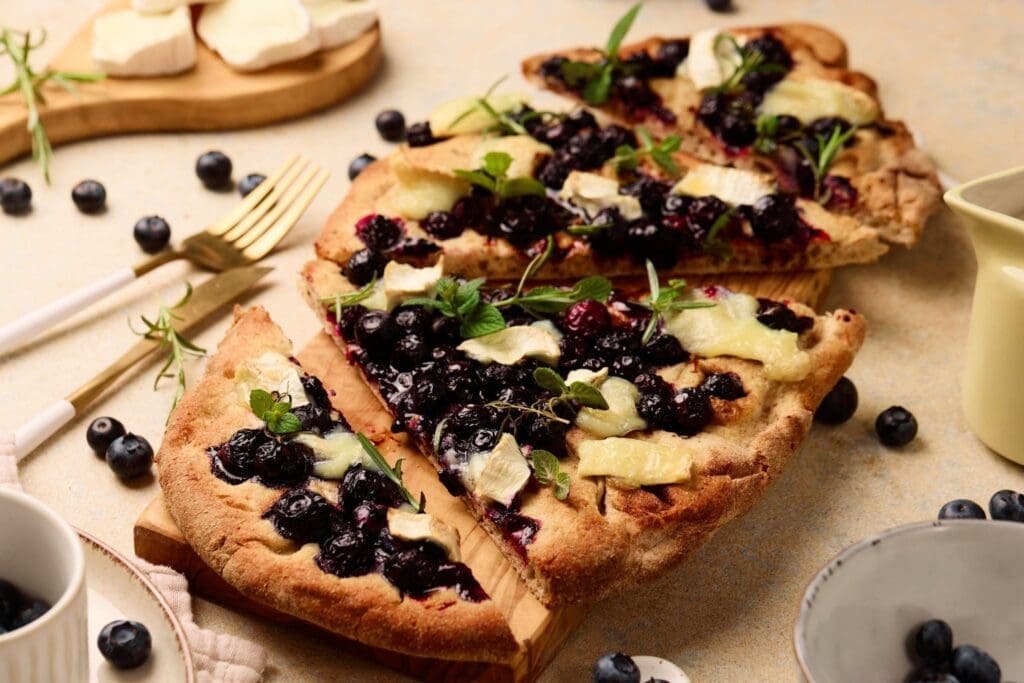 A rectangular blueberry goat cheese flatbread topped with melted cheese and fresh herbs, sliced on a wooden board. Blueberries, herbs, and cheese are scattered around, with plates and cutlery nearby.