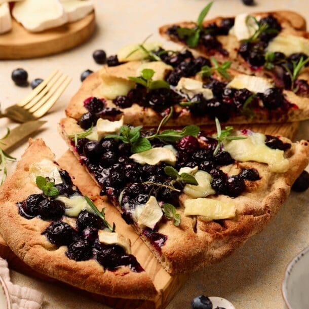 A rectangular blueberry goat cheese flatbread topped with melted cheese and fresh herbs, sliced on a wooden board. Blueberries, herbs, and cheese are scattered around, with plates and cutlery nearby.