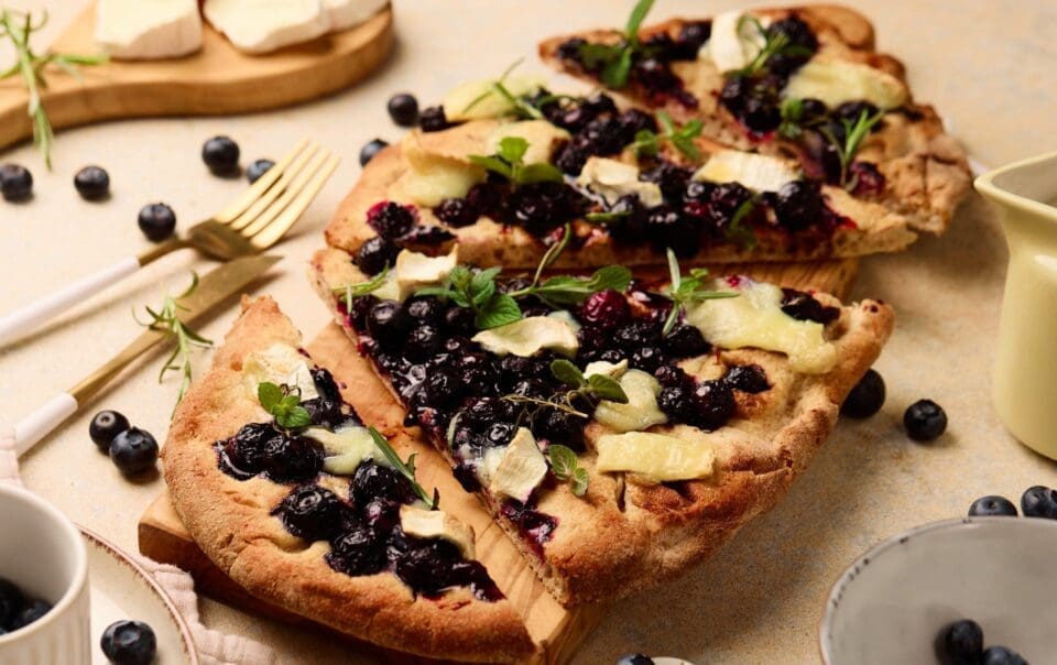 A rectangular blueberry goat cheese flatbread topped with melted cheese and fresh herbs, sliced on a wooden board. Blueberries, herbs, and cheese are scattered around, with plates and cutlery nearby.