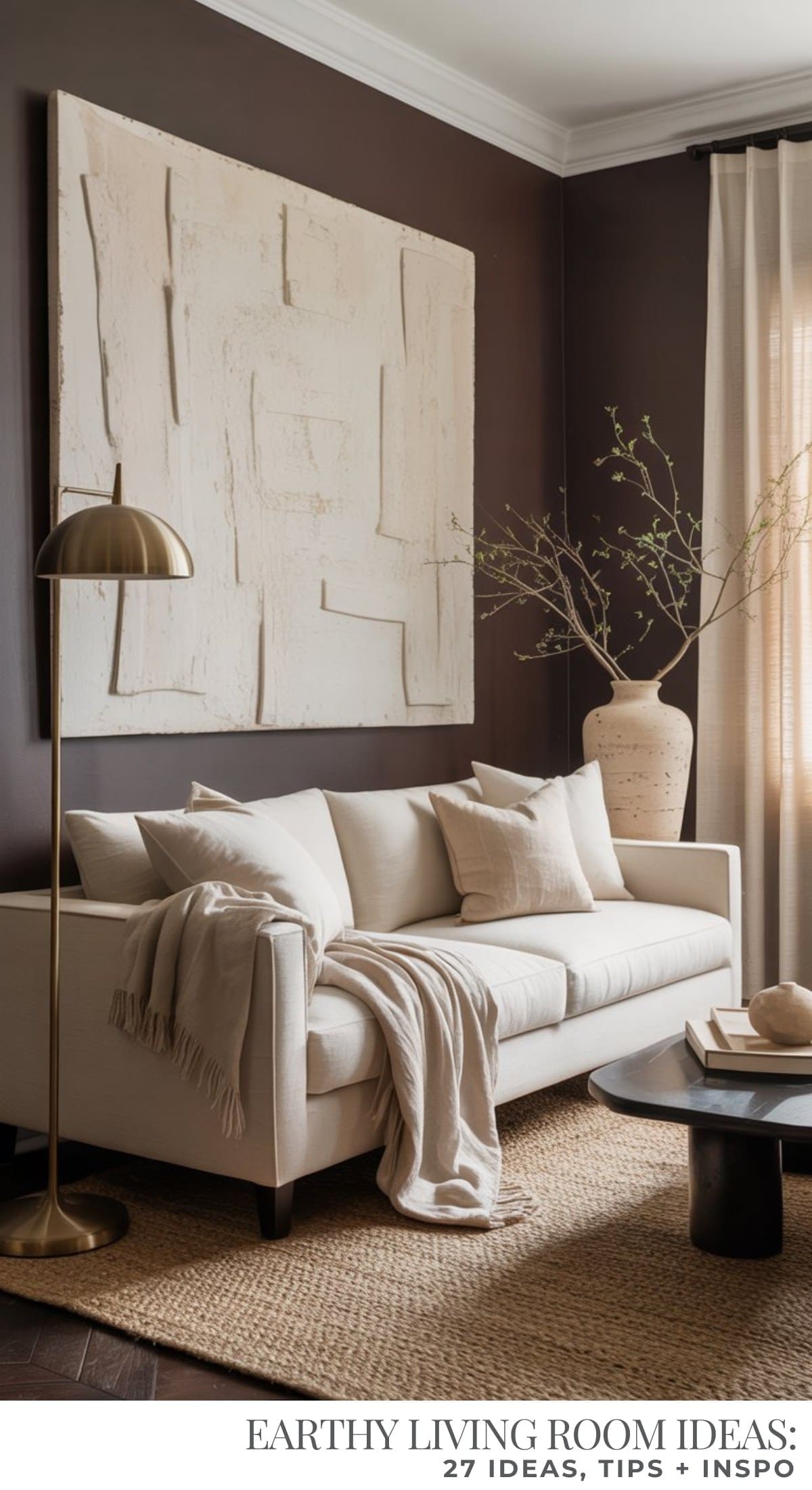 A modern living room with a cream sofa, neutral pillows, a beige throw, and a textured rug. Large abstract wall art and a tall vase with branches decorate the dark chocolate brown walls, complemented by a gold floor lamp.