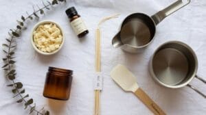 A Eucalyptus DIY Candle kit on a white cloth, featuring wax beads, a brown glass jar, fragrance oil, two metal saucepans, a spatula, wicks, and a fresh eucalyptus sprig all arranged neatly.