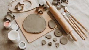 Air dry clay, cookie cutters, a rolling pin, paintbrushes, and small jars of paint arranged on a beige fabric surface, with a rolled-out piece of clay on a wooden board and clay shapes nearby.