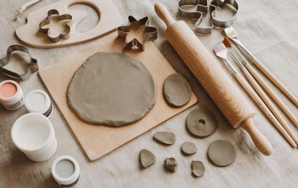 Air dry clay, cookie cutters, a rolling pin, paintbrushes, and small jars of paint arranged on a beige fabric surface, with a rolled-out piece of clay on a wooden board and clay shapes nearby.