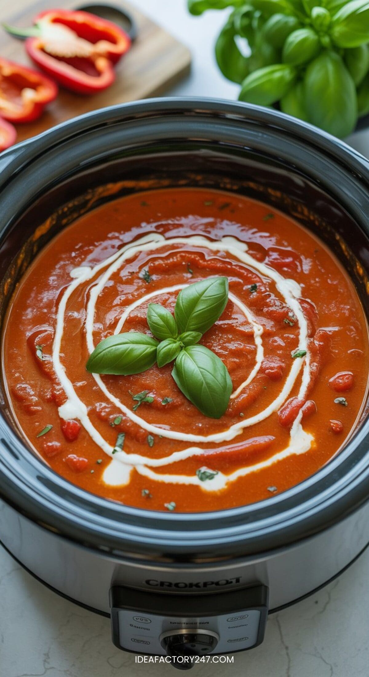 A crockpot soup recipe featuring creamy tomato soup in a slow cooker, topped with a swirl of cream and fresh basil. In the background, sliced red bell peppers and a bunch of basil add vibrant color to the countertop scene.