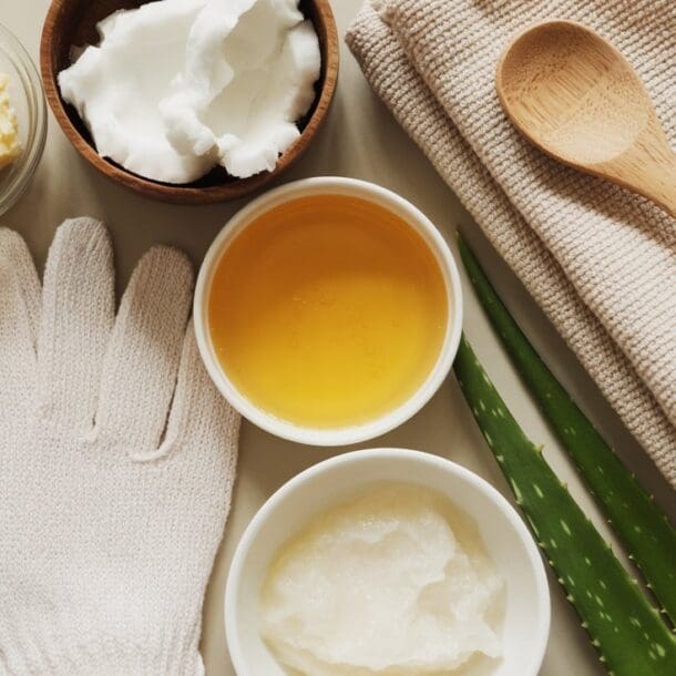 A flat lay of skincare ingredients and tools for a DIY hand mask, including shea butter, coconut oil, honey, scrub, two aloe vera leaves, a wooden spoon, beige towels, and a white exfoliating glove.