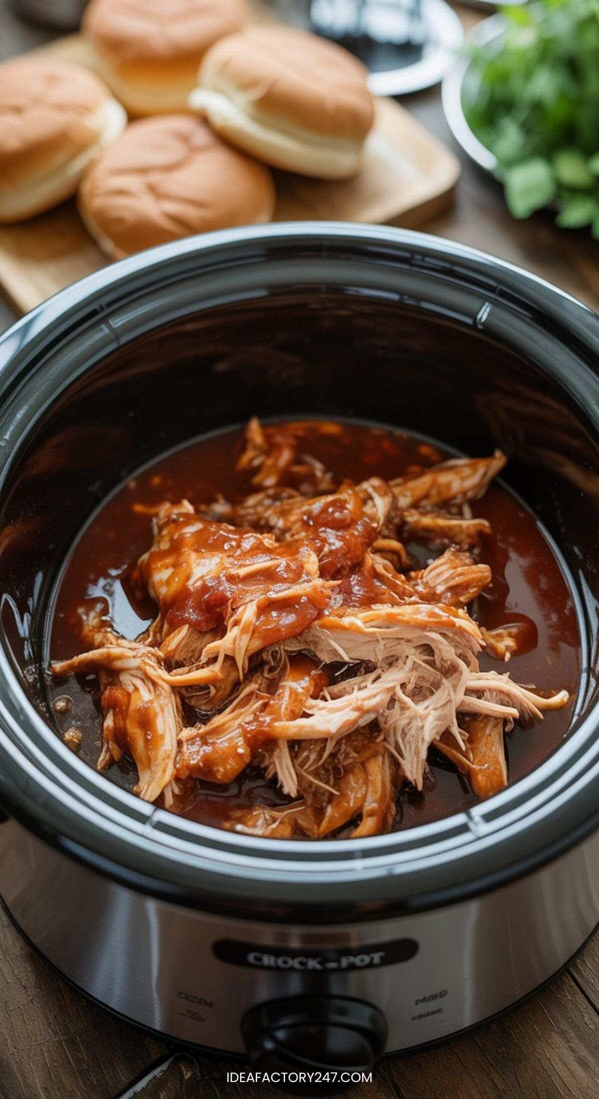 Shredded barbecue chicken in a slow cooker with sauce, ready to be served—an ideal choice for easy crockpot meals. In the background, hamburger buns and fresh greens are placed on a wooden table.