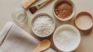 Flat lay of bowls with white and brown powders, a glass jar, a wooden spoon, two brushes, and a folded beige towel on a light surface, suggesting ingredients for homemade skincare or DIY beauty treatments.